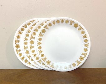 May include: A set of four round white dinner plates featuring a repeating gold floral pattern along the rim. The plates are stacked slightly offset, displaying the consistent circular design of stylized flowers and leaves on each piece.