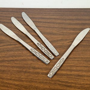 May include: Four stainless steel butter knives with a decorative floral pattern on the handles. The knives are arranged on a dark brown wooden surface. The blades are smooth and the handles have a detailed design.