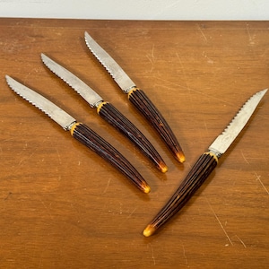 May include: Four steak knives with serrated blades and brown handles with yellow accents are arranged on a wooden surface. The knives have a classic design, suitable for dining.