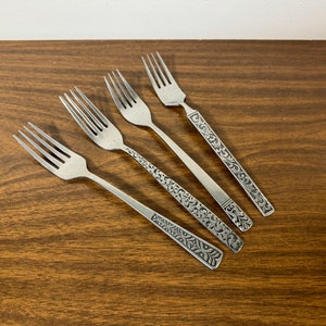 May include: Five silver forks with ornate, engraved handles are arranged diagonally on a dark wood surface. The forks have four tines each and a decorative pattern on the handle. The background is a plain white wall.