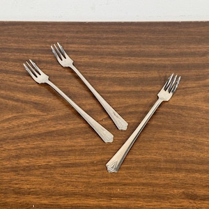 May include: Three silver forks with decorative handles are arranged on a dark brown wooden surface. Each fork has three tines. The forks are angled in different directions, creating a dynamic composition.
