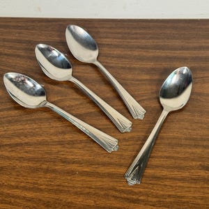 May include: Four silver spoons with a decorative handle design are arranged on a dark brown wooden surface. The spoons are positioned in a slightly scattered pattern, reflecting light.