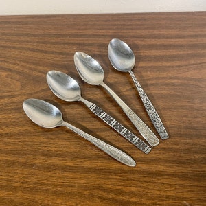 May include: Four silver spoons with different handle designs are arranged on a brown wooden surface. The spoons have oval bowls and ornate patterns on their handles, including floral and geometric motifs. The spoons are likely for eating.