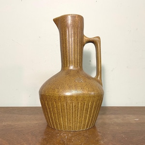 May include: A brown ceramic pitcher with a speckled finish, featuring a wide, rounded base with vertical fluted detailing, a tall, slender neck with a matching fluted design, a small pouring spout, and a single integrated side handle.