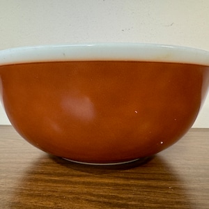 May include: A large, brown glass mixing bowl with a white rim. The bowl is sitting on a wooden surface.