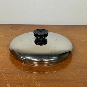 May include: A circular, polished stainless steel pot lid featuring a smooth, reflective surface and a centered, black, cylindrical knob handle. The lid has a slightly domed shape with a flat rim designed to fit securely onto a matching cooking vessel.