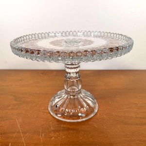 May include: A clear glass cake stand with a round, beaded edge. The stand has a decorative stem and base. The cake stand is approximately 10 inches in diameter and is suitable for displaying desserts.