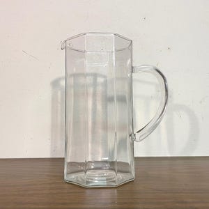 May include: A clear glass pitcher featuring a distinct octagonal shape with flat vertical panels. The vessel includes a curved handle attached to one side and a small pouring spout at the top rim, showcasing a transparent and smooth glass construction.