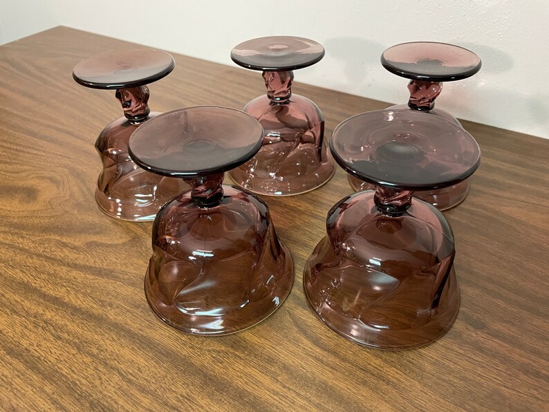 May include: Five clear, amethyst-colored glass dessert dishes with a swirl design. The dishes have a footed base.