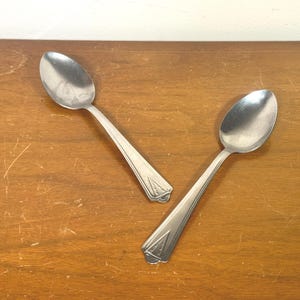 May include: Two silver-toned metal spoons with a polished finish. Each spoon features a tapered handle with a subtle, engraved triangular geometric pattern near the base. The bowls of the spoons are oval-shaped and smooth, reflecting light on their surface.