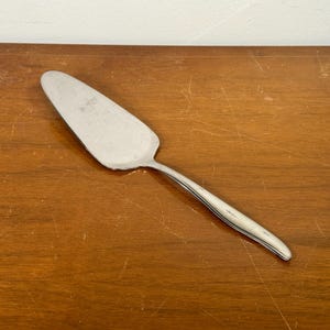 May include: A silver pie server with a triangular blade and a long, slender handle. The server is made of stainless steel and is designed for serving pies, cakes, or other baked goods. The server is resting on a wooden surface.