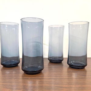 May include: A set of four tall drinking glasses made of translucent blue glass. Each glass features a cylindrical shape with a slightly wider top and a thicker, darker blue base. The glass color transitions from a deep navy at the bottom to a lighter, soft blue hue toward the rim.