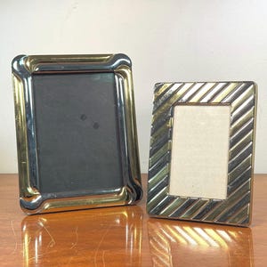 Peut inclure: Deux cadres photo rectangulaires. L'un a une bordure dorée et argentée avec un intérieur noir. L'autre a une bordure à rayures diagonales dorées et argentées avec un intérieur blanc. Les deux cadres sont sur une surface en bois.