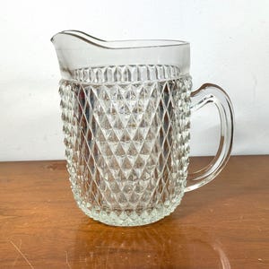 May include: Clear glass pitcher with a diamond pattern. The pitcher has a handle and a spout for pouring. It is sitting on a wooden surface. The pitcher is a vintage item.