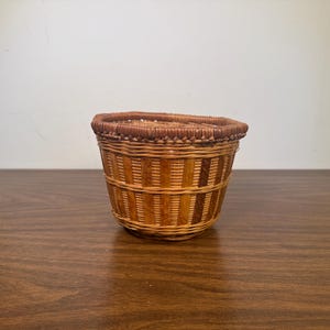 May include: A small, woven basket with a rounded shape. The basket is made of natural-colored reeds and features vertical stripes of darker brown reeds. The rim is a darker brown color. The basket sits on a wooden surface.