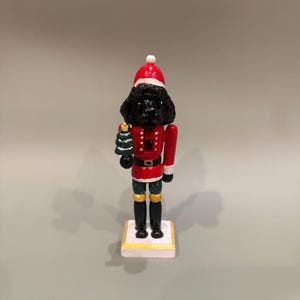 May include: A decorative Christmas nutcracker in the shape of a black dog wearing a red Santa suit and hat. The dog holds a small green Christmas tree. The nutcracker stands on a white base with gold trim.