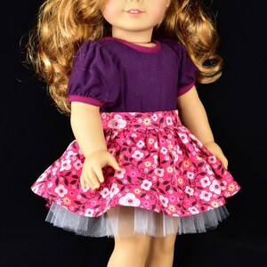K&ouml;nnte beinhalten: Eine Puppe mit einem lila Oberteil und einem rosa Blumenrock mit einem wei&szlig;en T&uuml;ll-Unterrock. Die Puppe hat welliges, rotbraunes Haar und ein rotes Blumen-Haaraccessoire. Die Puppe tr&auml;gt schwarze Glitzerschuhe.
