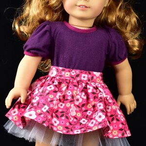 K&ouml;nnte beinhalten: Puppe mit langem, gewelltem blondem Haar und blauen Augen, tr&auml;gt ein lila Oberteil mit kurzen &Auml;rmeln und einen rosa Blumenrock mit wei&szlig;em T&uuml;llunterrock. Eine rote Blume schm&uuml;ckt das Haar der Puppe. Die Puppe steht vor schwarzem Hintergrund.