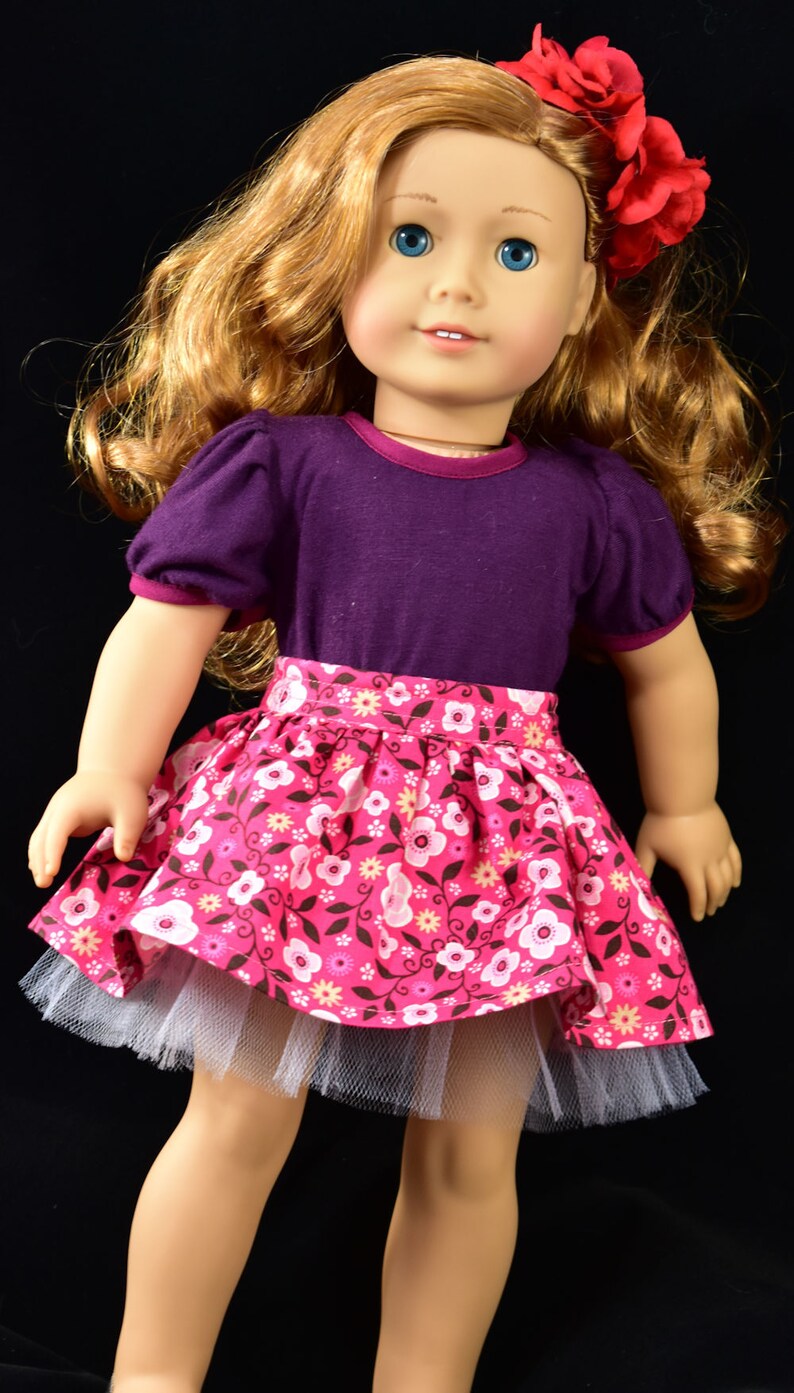 K&ouml;nnte beinhalten: Puppe mit langem, gewelltem blondem Haar und blauen Augen, tr&auml;gt ein lila Oberteil mit kurzen &Auml;rmeln und einen rosa Blumenrock mit wei&szlig;em T&uuml;llunterrock. Eine rote Blume schm&uuml;ckt das Haar der Puppe. Die Puppe steht vor schwarzem Hintergrund.