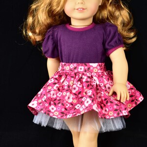 K&ouml;nnte beinhalten: Puppe mit blonden Haaren und blauen Augen, tr&auml;gt ein lila Oberteil und einen rosa Blumenrock. Der Rock hat einen wei&szlig;en T&uuml;ll-Unterrock. Die Puppe hat eine rote Blume im Haar und tr&auml;gt schwarze Schuhe.