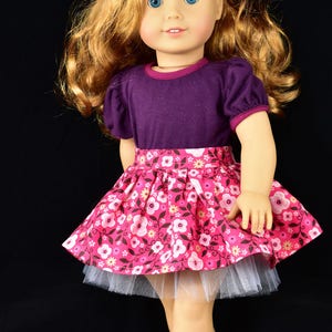 K&ouml;nnte beinhalten: Puppe mit einem lila kurz&auml;rmeligen Oberteil und einem rosa Blumenrock sowie schwarzen Schuhen. Der Rock hat einen wei&szlig;en T&uuml;ll-Unterrock. Die Puppe hat blondes Haar und ein rotes Blumen-Haaraccessoire.