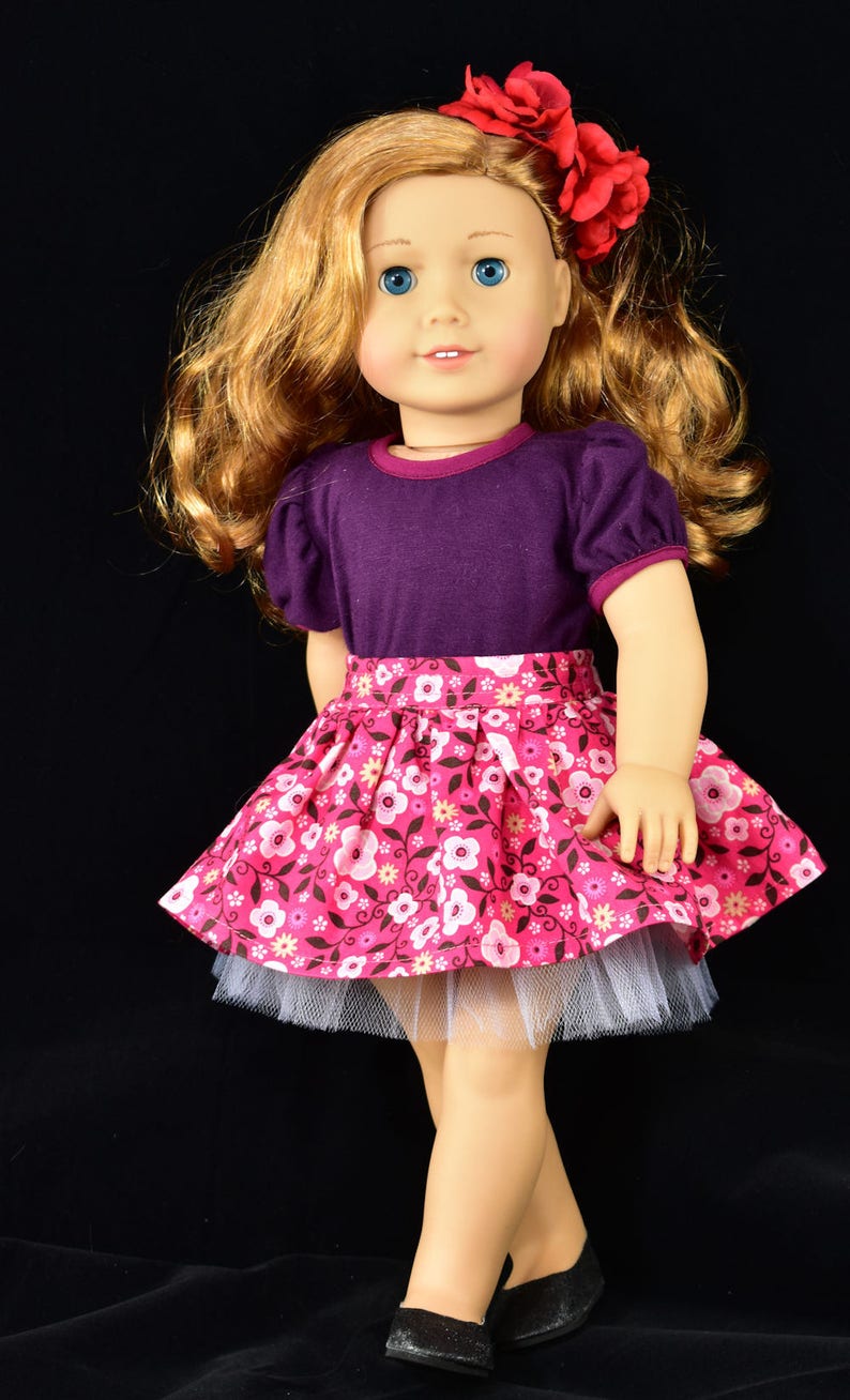 K&ouml;nnte beinhalten: Puppe mit einem lila kurz&auml;rmeligen Oberteil und einem rosa Blumenrock sowie schwarzen Schuhen. Der Rock hat einen wei&szlig;en T&uuml;ll-Unterrock. Die Puppe hat blondes Haar und ein rotes Blumen-Haaraccessoire.