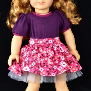 K&ouml;nnte beinhalten: Puppe mit blonden Haaren und blauen Augen, bekleidet mit einem lila Oberteil, einem rosa Blumenrock mit wei&szlig;em T&uuml;llunterrock und schwarzen Schuhen. Eine rote Blume schm&uuml;ckt das Haar der Puppe. Kinderspielzeug.
