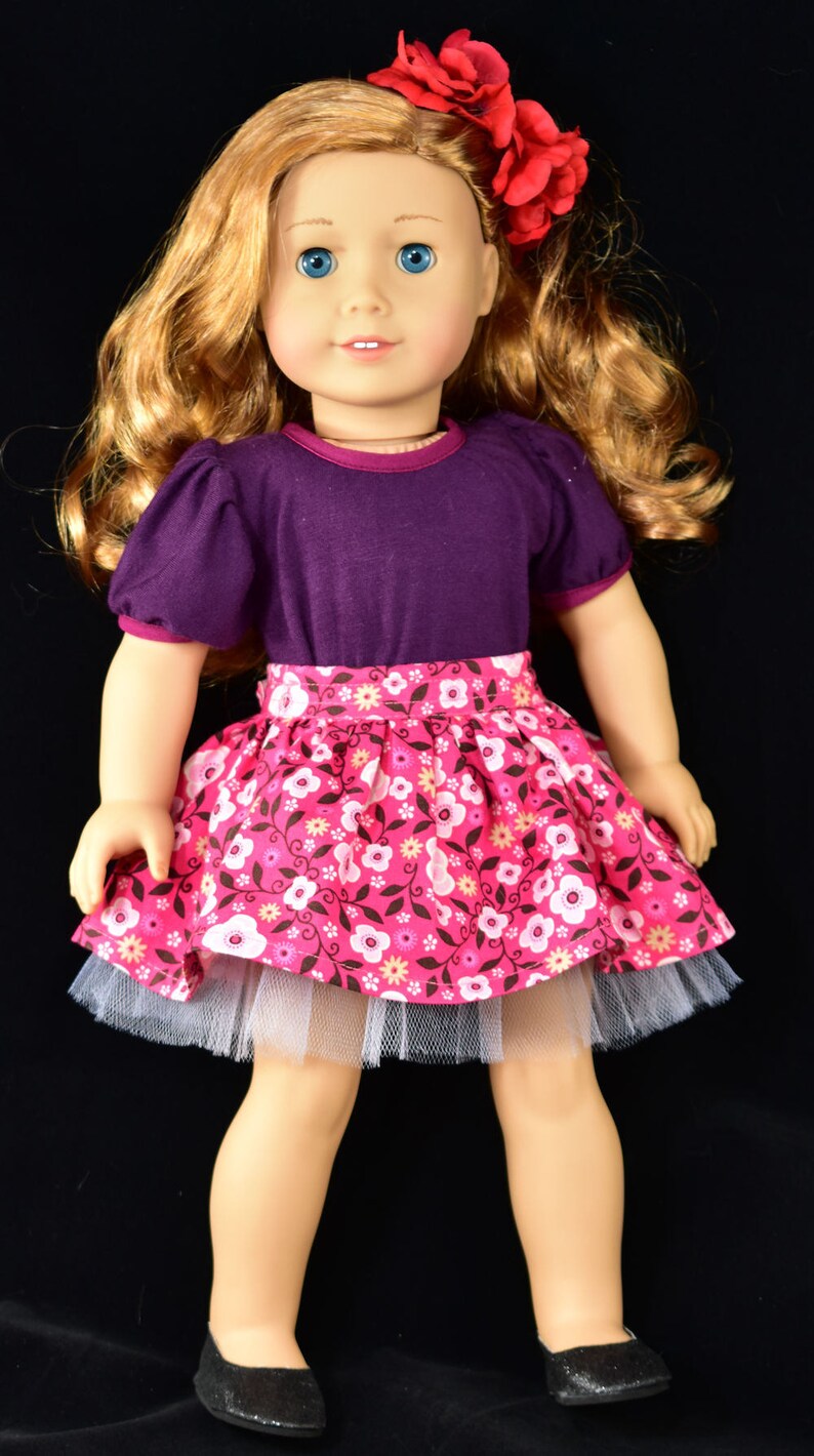 K&ouml;nnte beinhalten: Puppe mit blonden Haaren und blauen Augen, bekleidet mit einem lila Oberteil, einem rosa Blumenrock mit wei&szlig;em T&uuml;llunterrock und schwarzen Schuhen. Eine rote Blume schm&uuml;ckt das Haar der Puppe. Kinderspielzeug.