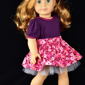 K&ouml;nnte beinhalten: Puppe mit langem, gewelltem blondem Haar und blauen Augen, tr&auml;gt ein lila Oberteil und einen rosa Blumenrock sowie schwarze Glitzerschuhe. Eine rote Blume schm&uuml;ckt das Haar der Puppe.