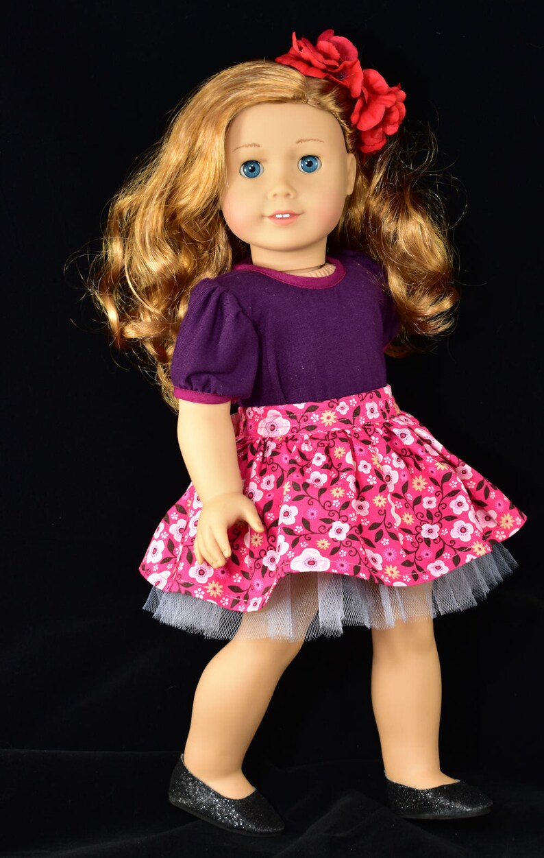 K&ouml;nnte beinhalten: Puppe mit langem, gewelltem blondem Haar und blauen Augen, tr&auml;gt ein lila Oberteil und einen rosa Blumenrock sowie schwarze Glitzerschuhe. Eine rote Blume schm&uuml;ckt das Haar der Puppe.