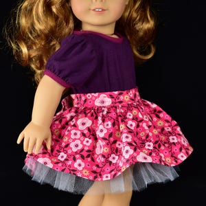 K&ouml;nnte beinhalten: Puppe mit einem lila Oberteil mit kurzen &Auml;rmeln und einem rosa Blumenrock mit wei&szlig;em T&uuml;llunterrock. Die Puppe hat lange, wellige, rotbraune Haare, blaue Augen und eine rote Blume als Haaraccessoire. Schwarze Glitzer-Ballerinas vervollst&auml;ndigen das Outfit.