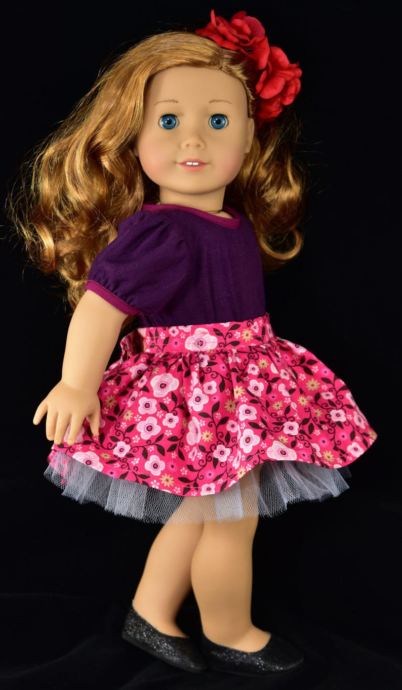 K&ouml;nnte beinhalten: Puppe mit einem lila Oberteil mit kurzen &Auml;rmeln und einem rosa Blumenrock mit wei&szlig;em T&uuml;llunterrock. Die Puppe hat lange, wellige, rotbraune Haare, blaue Augen und eine rote Blume als Haaraccessoire. Schwarze Glitzer-Ballerinas vervollst&auml;ndigen das Outfit.