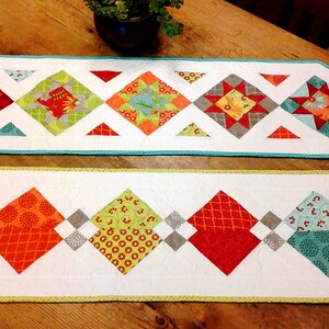 May include: Two decorative table runners with a white background and colourful diamond patterns. The diamonds feature red, orange, green, and grey designs. The edges are trimmed with teal and yellow borders. The runners are placed on a wooden table.