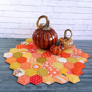 May include: A handmade, hexagonal patchwork table runner in shades of orange, yellow, and green, with a variety of fabric patterns. Three glass pumpkins in varying sizes and shades of orange sit atop the runner, creating a fall-themed display.