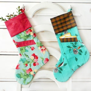 May include: Two handmade Christmas stockings. One has a light blue radish pattern and a red cuff. The other has a teal bird and pinecone design and a brown plaid cuff. Both have a pocket. The stockings are hanging on a white chair.