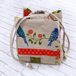 Pode incluir: Uma bolsa bege com cordão com um design de pássaros e flores. A bolsa apresenta dois pássaros azuis empoleirados em um galho com rosas vermelhas. Uma faixa vermelha com bolinhas laranja adorna a frente. A bolsa tem cordões bege e contas de madeira.