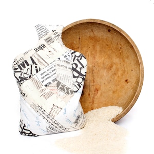 Può includere: Una borsa di stoffa bianca con testo bianco e nero stampato su di essa, seduta su una ciotola di legno piena di riso bianco. La borsa sta versando riso su una superficie bianca.