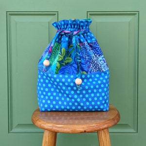 May include: A blue and floral drawstring bag with a polka dot bottom. The bag has wooden toggles and is sitting on a wooden stool.