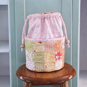 May include: A patchwork fabric drawstring bag with a floral and geometric pattern in shades of yellow, orange, and green. The top section is a pink fabric with printed text. The bag has a wooden bead on each drawstring.