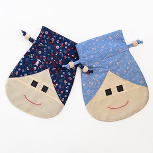 May include: Two fabric drawstring bags with a whimsical face design. One bag is navy blue with a floral pattern, and the other is light blue with a similar floral print. Both have beige faces, black square eyes, and red stitched smiles.