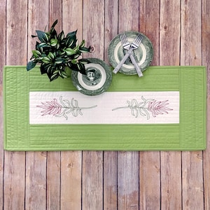May include: A green quilted table runner with a white central panel featuring embroidered leaf designs in green and burgundy. Two decorative plates with forks and a small plant are on the runner, set against a wooden background.