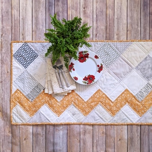 May include: A quilted table runner with a geometric design in cream, grey, and orange. A white plate with red floral accents, silverware, a fern, and napkins are arranged on the runner, creating a rustic table setting.