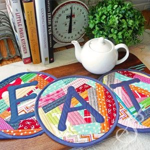 Op de afbeelding: Drie kleurrijke stoffen pottenlapjes met de letters "EAT" in blauwe stof erop. De pottenlapjes liggen op een houten oppervlak met een witte theepot en een vintage weegschaal op de achtergrond.