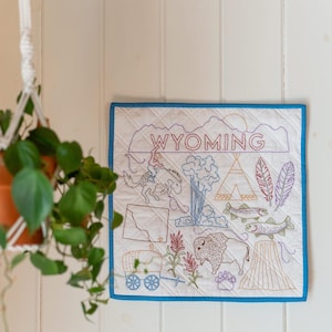 May include: A quilted wall hanging featuring an embroidered map of Wyoming. The white fabric has a blue border and is decorated with images of a cowboy, bison, teepee, and the word "WYOMING".