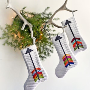 May include: Three white Christmas stockings with black arrow designs and colorful fabric accents hanging on a white antler.