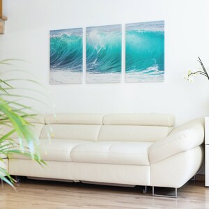 May include: A white leather sofa with a white wall behind it. There is a three-piece canvas print of a blue ocean wave on the wall. A potted plant is in the foreground.