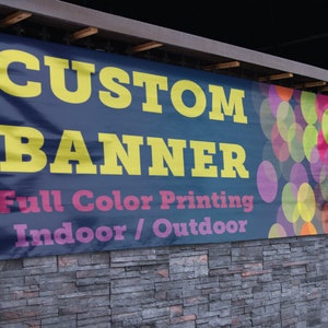 May include: A blue banner with the text "CUSTOM BANNER" in yellow letters. The banner also says "Full Color Printing Indoor/Outdoor" in pink and white letters. The banner has a colorful background of overlapping circles.