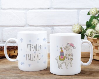 May include: Two white ceramic mugs with different designs. One mug features the text "LITERALLY FREEZING" with blue snowflakes. The other mug features a cartoon llama wearing a Santa hat and carrying presents.