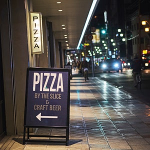 May include: A blue and white sign with the text "Pizza by the Slice & Craft Beer" and an arrow pointing to the left. The sign is on a sidewalk in front of a building with a lit sign that says "Pizza".