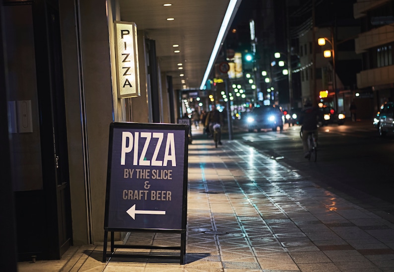 May include: A blue and white sign with the text "Pizza by the Slice & Craft Beer" and an arrow pointing to the left. The sign is on a sidewalk in front of a building with a lit sign that says "Pizza".